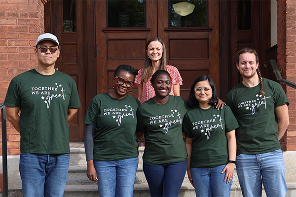 A group of students stands outside a red brick building. They are wearing green shirts that read, "Together we are GREAT."