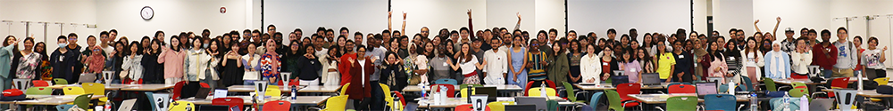 A large group of students stands in a line at the end of a conference room, smiling and gesturing at the camera.