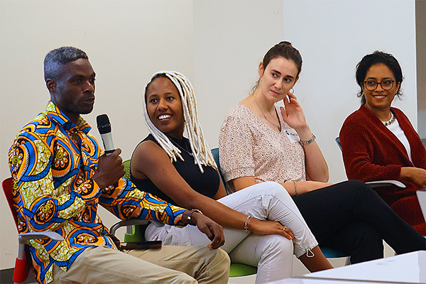 A man in a yellow and blue shirt is speaking into a microphone while seated. A group of panelists watch and listen to him.