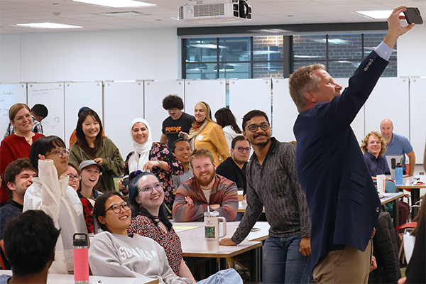 President Guskiewicz standing at the front of a large conference room, taking a photo of himself with a group of students seated behind him.
