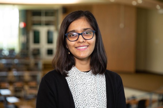 Lamia Bagasrawala in a black and white dotted shirt and black glasses, standing at the front of a classroom.