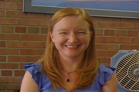 Betsy in a blue tank top, standing in front of a brick wall.