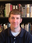 Matthew Yalch in a black shirt, standing in front of a bookshelf.