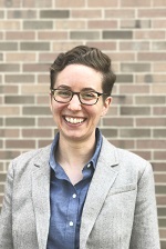 Mel Medeiros in a blue shirt and white overcoat, standing in front of a brick wall.