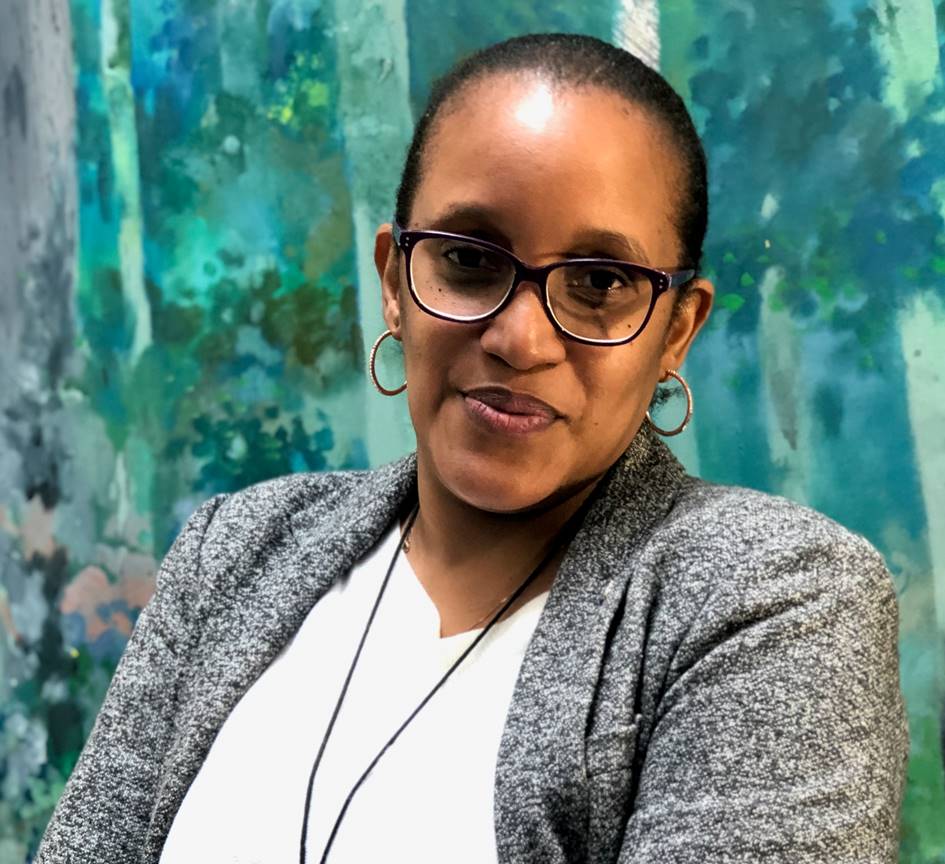 Angela Manjichi in a white undershirt and grey jacket, standing in front of a window.
