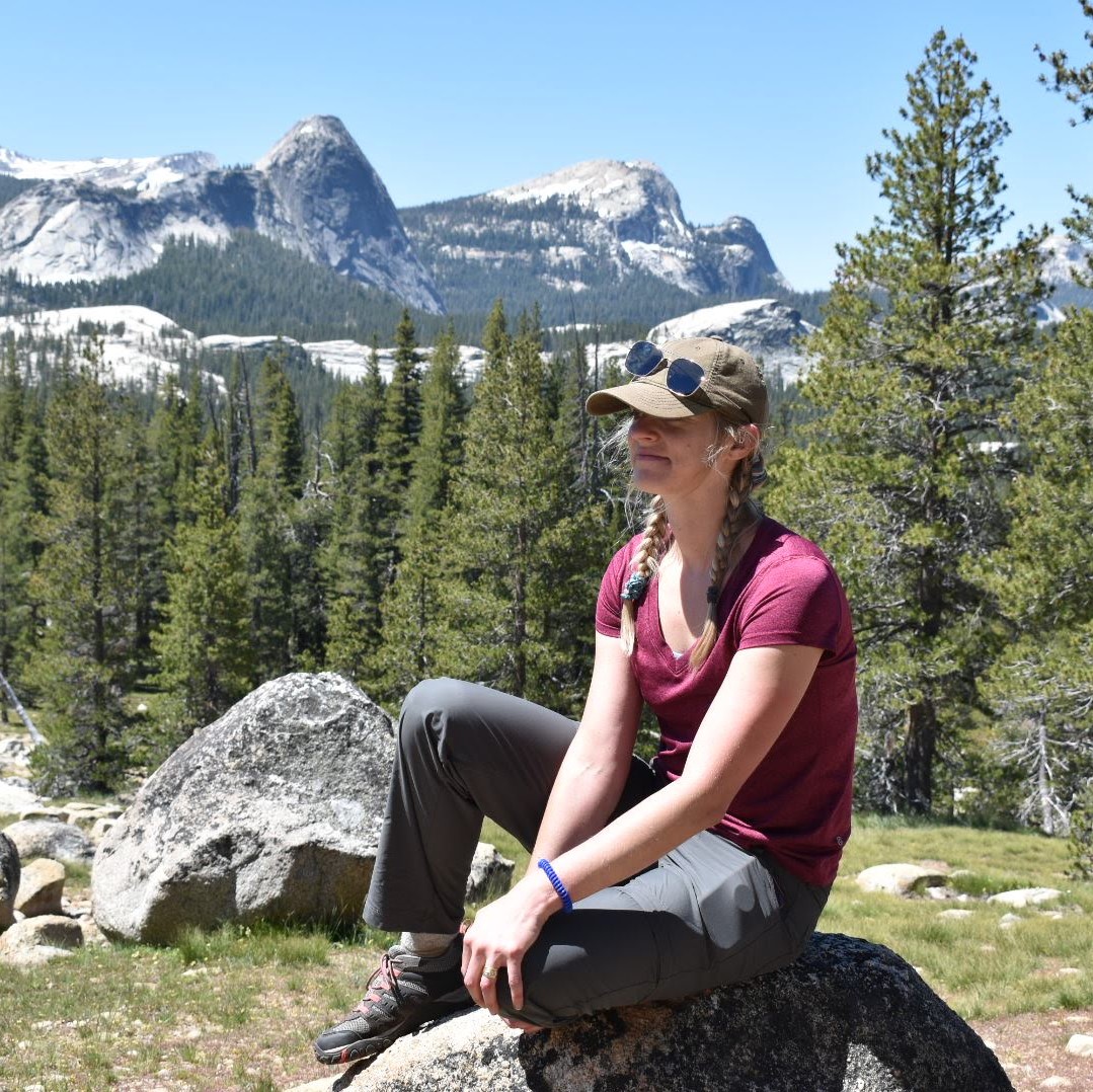 Christine Bennett in a red t-shirt and green hat, seated on a rock on a forested, mountainous landscape.