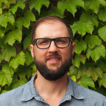 Ross Greedy in a blue shirt, standing in front of a wall of leaves.