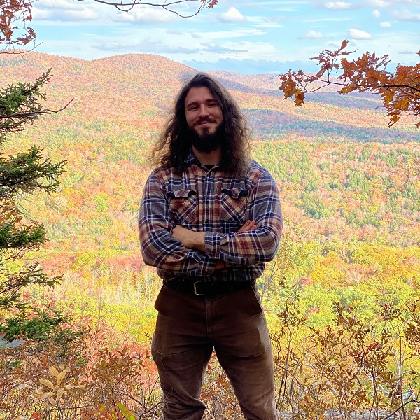 Blake Ginsburg in a brown and red plaid shirt, standing outside in front of a mountainous, wooded backdrop.