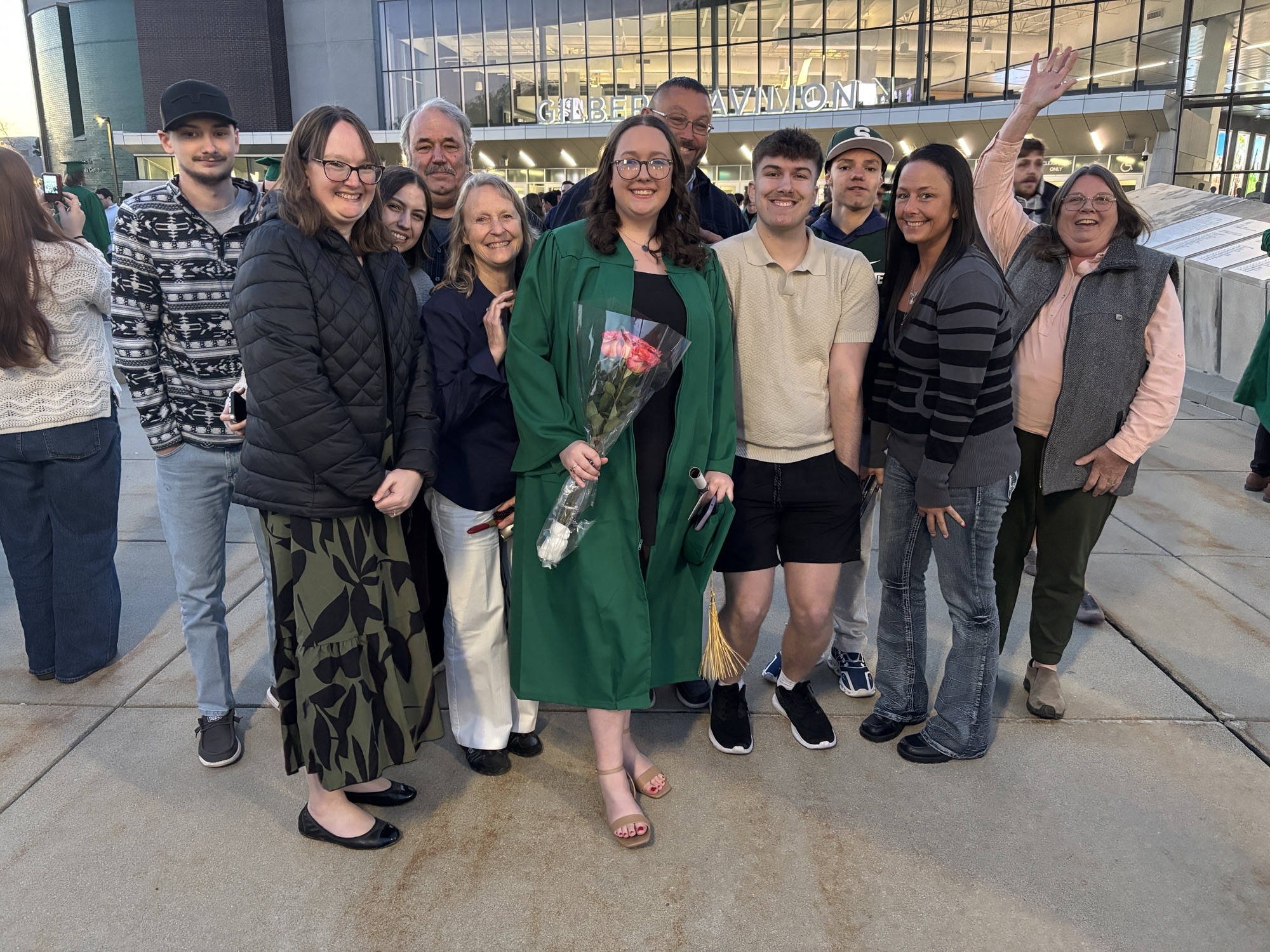 A group of about a dozen people stand together outside the Breslin Center at Michigan State University, smiling and posing with a graduate in a green cap and gown at the center, who is holding a bouquet of flowers and a rolled diploma.