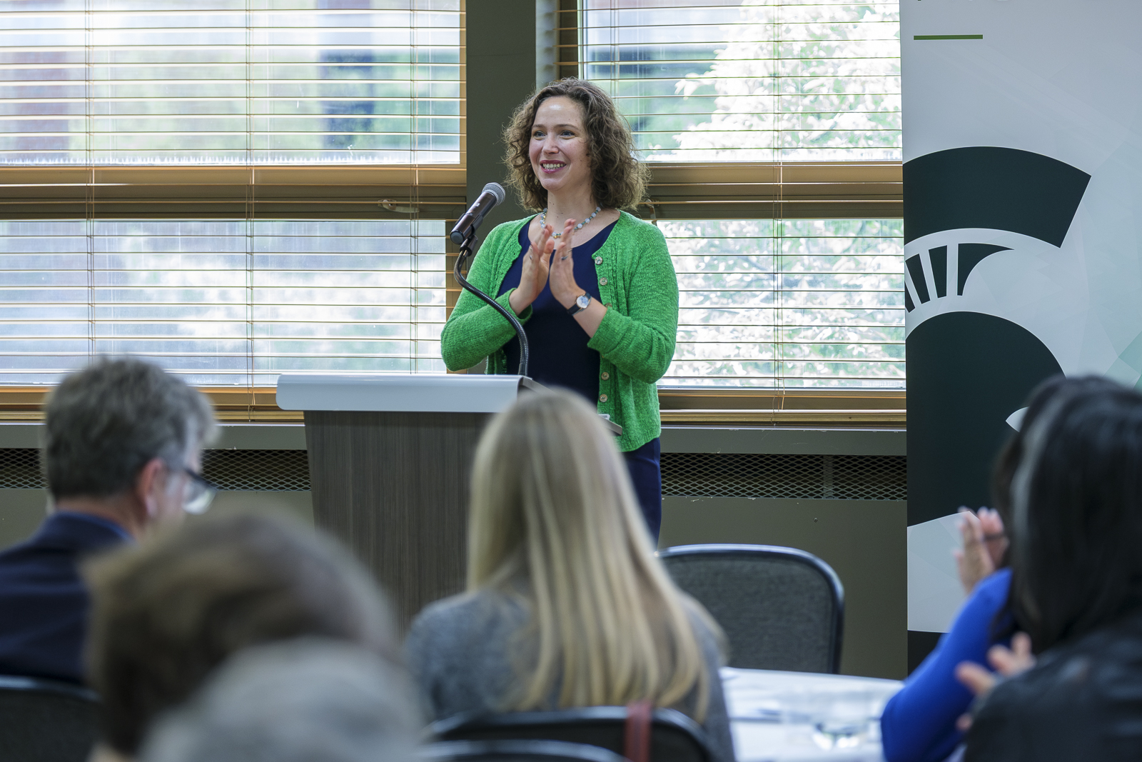 Professor Emily Conroy-Krutz stands at a podium and speaks to attendees during an MSU Department of History event. Audience members are seated in the foreground, with large windows and an MSU banner in the background.