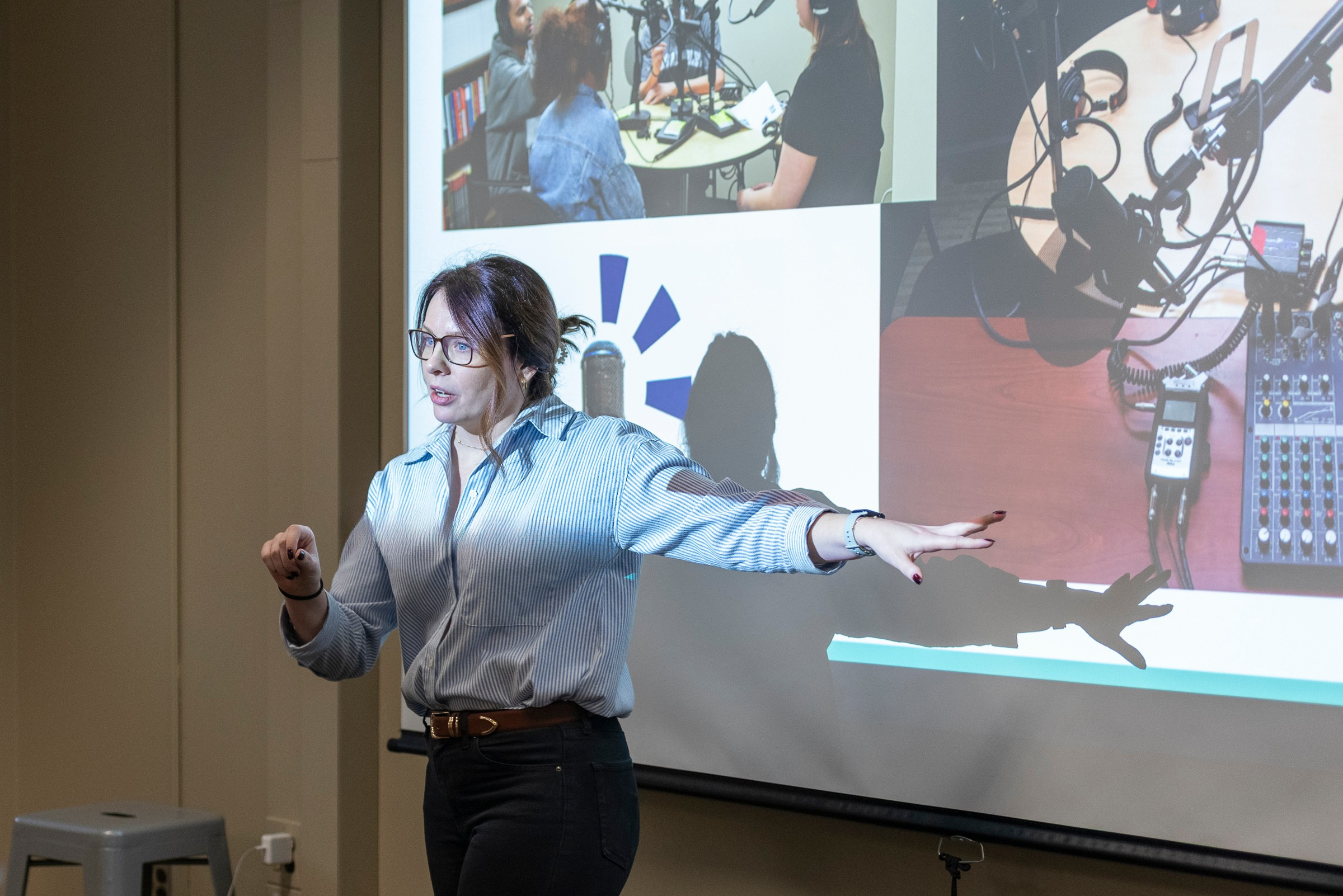 Gillian Macdonald gestures while speaking in front of a projected slide showing podcast recording equipment and students in a recording session.