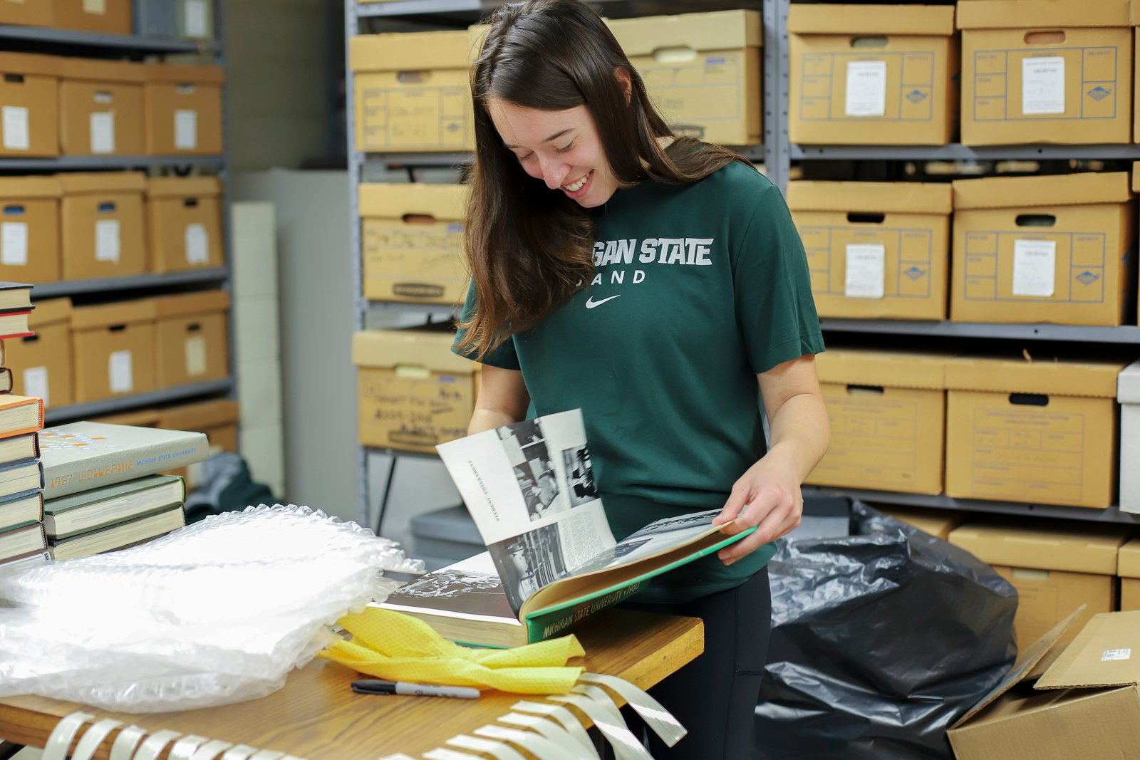 Student intern examines a bound archival volume in a storage area filled with shelves of archival boxes and preservation materials.