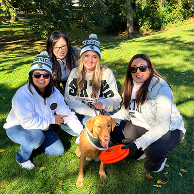 Four women dressed in Michigan State University apparel pose on a grassy hill with Zeke the Wonderdog
