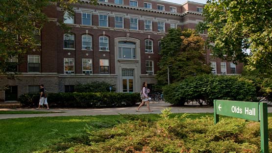 A photo of Olds Hall. Two students walk in front of the building. The trees are just starting to turn to fall colors. 