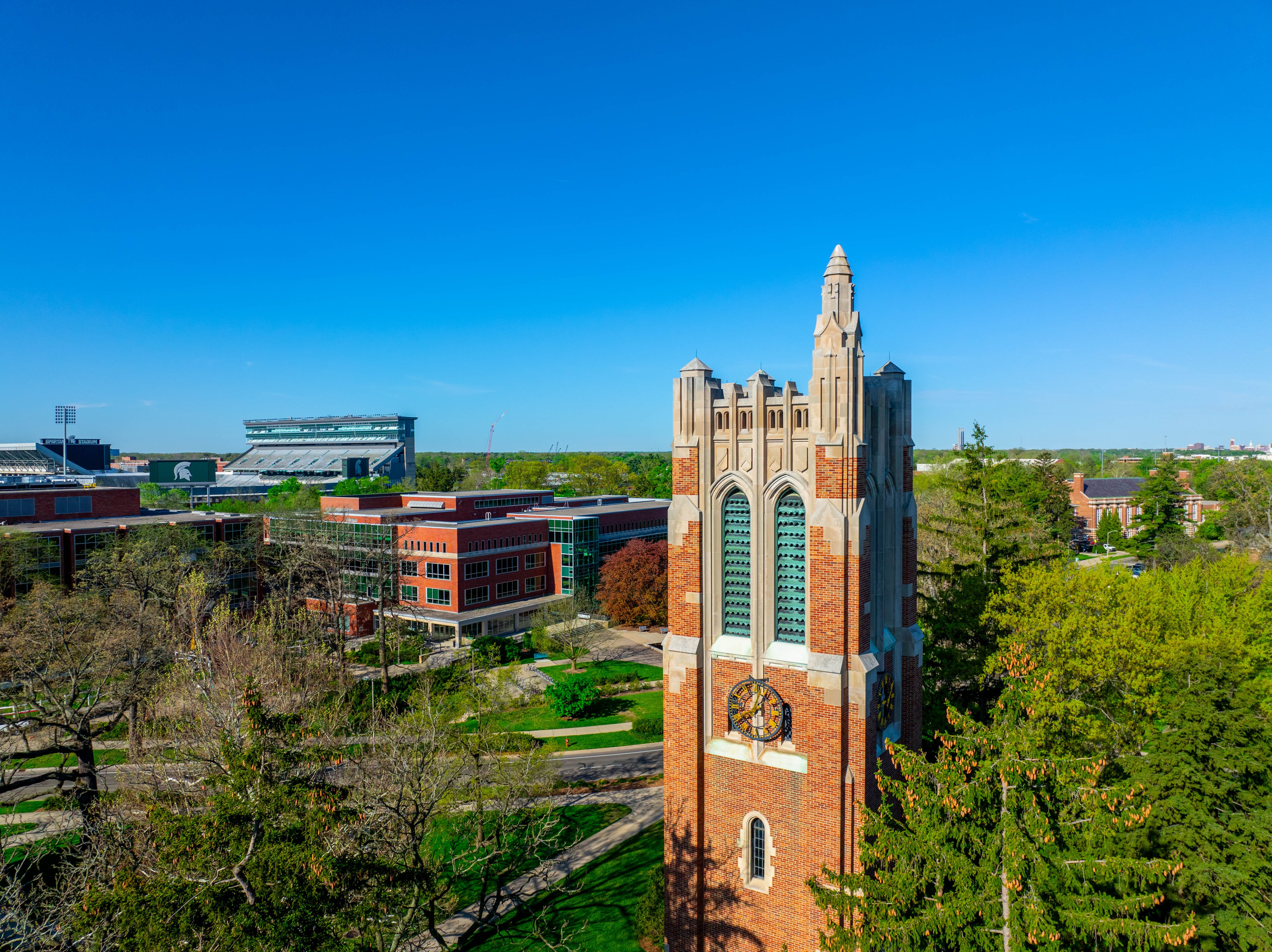 An aerial view of Beaumont Tower. The STEM Building and Spartan Stadium are visible in the background. The sky is a bright blue and free of clouds, and the surrounding trees are a bright spring green. 