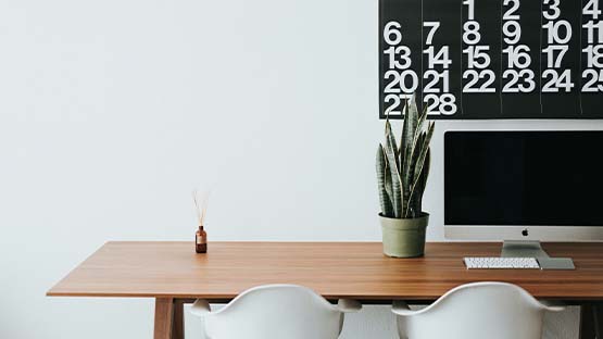 A wooden desk with two white desk chairs. There is a small plant, a computer monitor and a reed diffuser on the table. A black calendar with large white numbers hangs on the wall behind the computer.