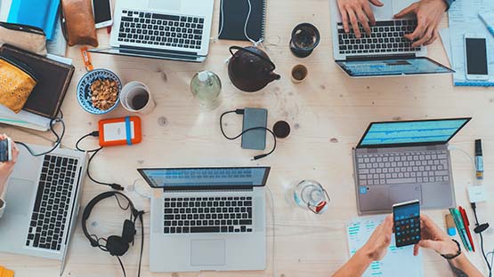 A view looking down on a collaborative workspace showing two pairs of hands, one holding a phone and one typing on a laptop. Two other laptops, a notebook and a pot of tea sit on the table.