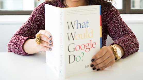 A woman sits at a table, reading the book "What Would Google Do"