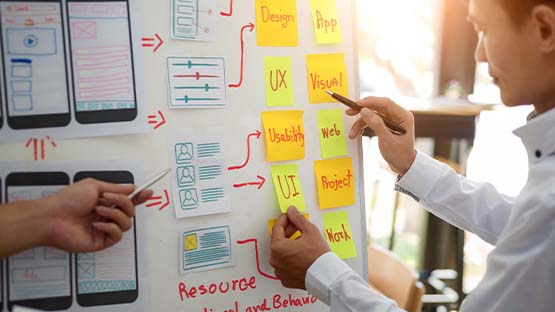 Two individuals stand in front of a white board. The whiteboard is covered with sticky notes labeled with marketing terms and wireframe mockups for a website.