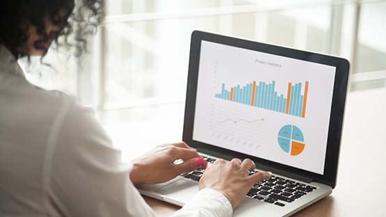 A photo of a woman sitting at a laptop with orange and turquoise graphs displayed on the screen. 