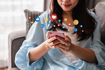 A woman sits in a chair, holding a phone in front of her face. Various social media reaction icons float out of the screen.