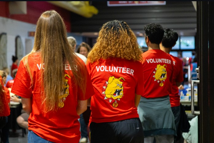 Three people wearing red volunteer shirts with a bulldog graphic stand together in a busy indoor setting.