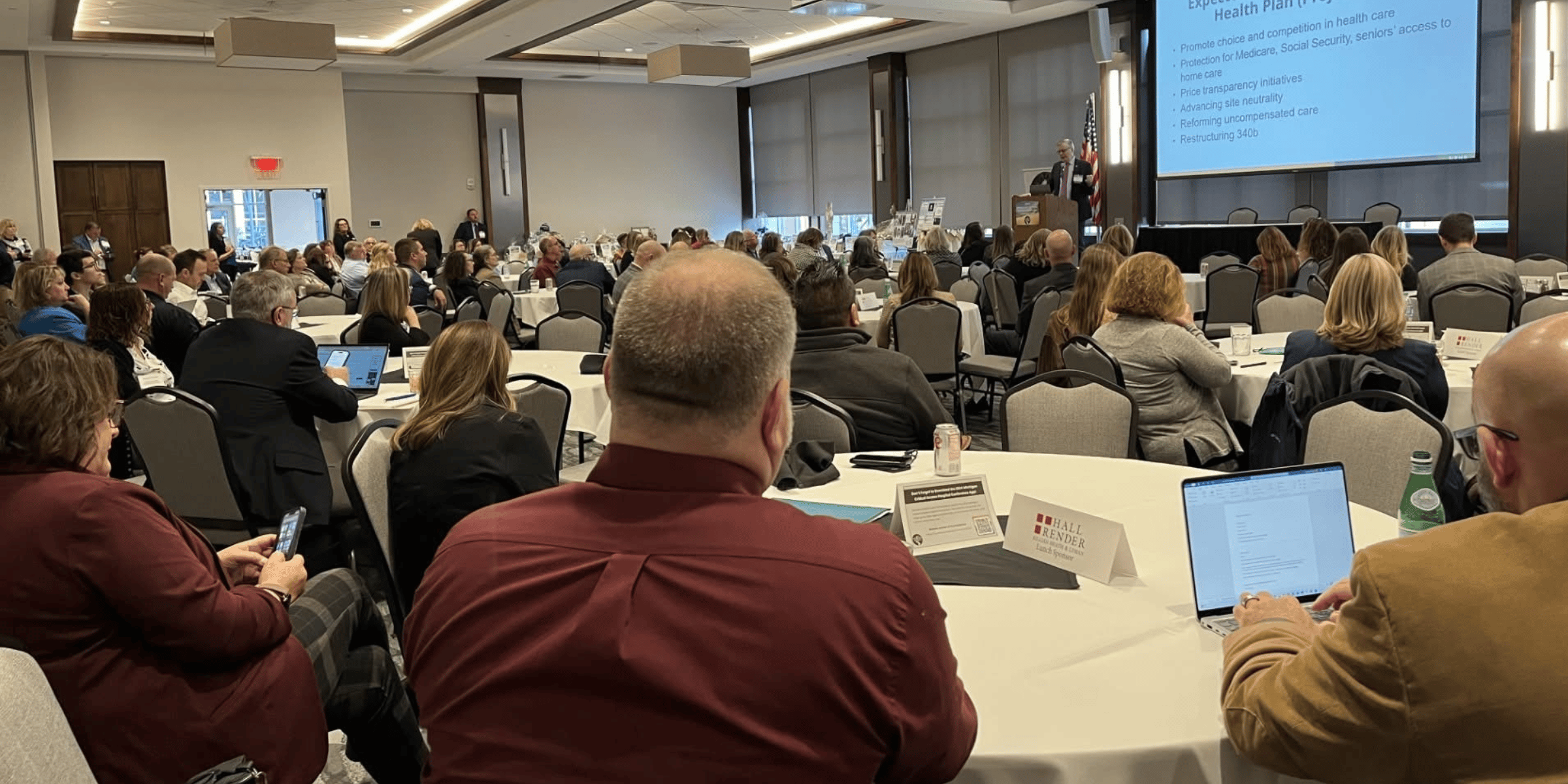 Conference attendees seated at round tables in a room, listening to a speaker at a podium with a presentation on a screen.