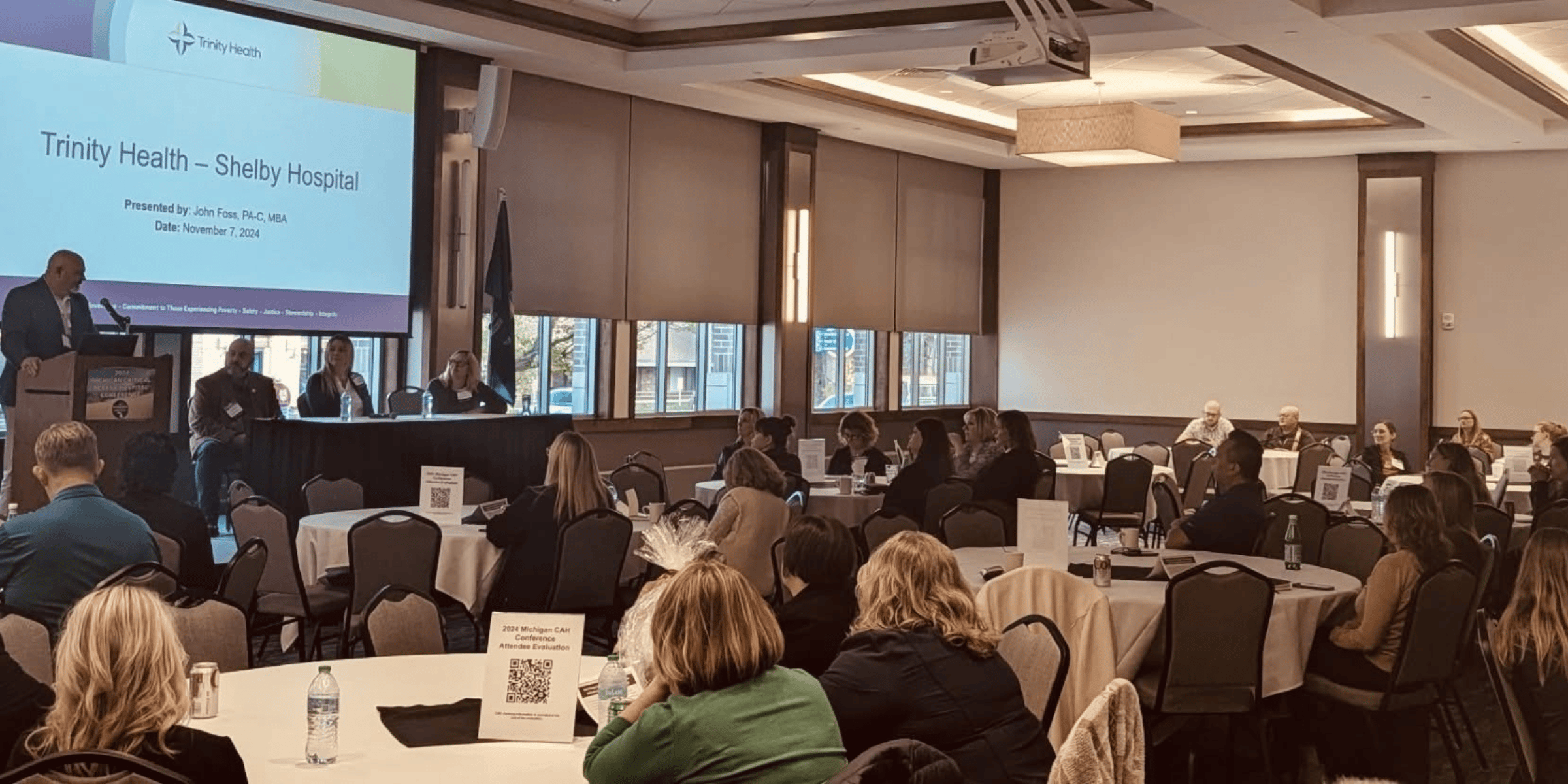 A conference room with a speaker at a podium in front of a screen displaying "Trinity Health – Shelby Hospital." Attendees are seated at round tables.