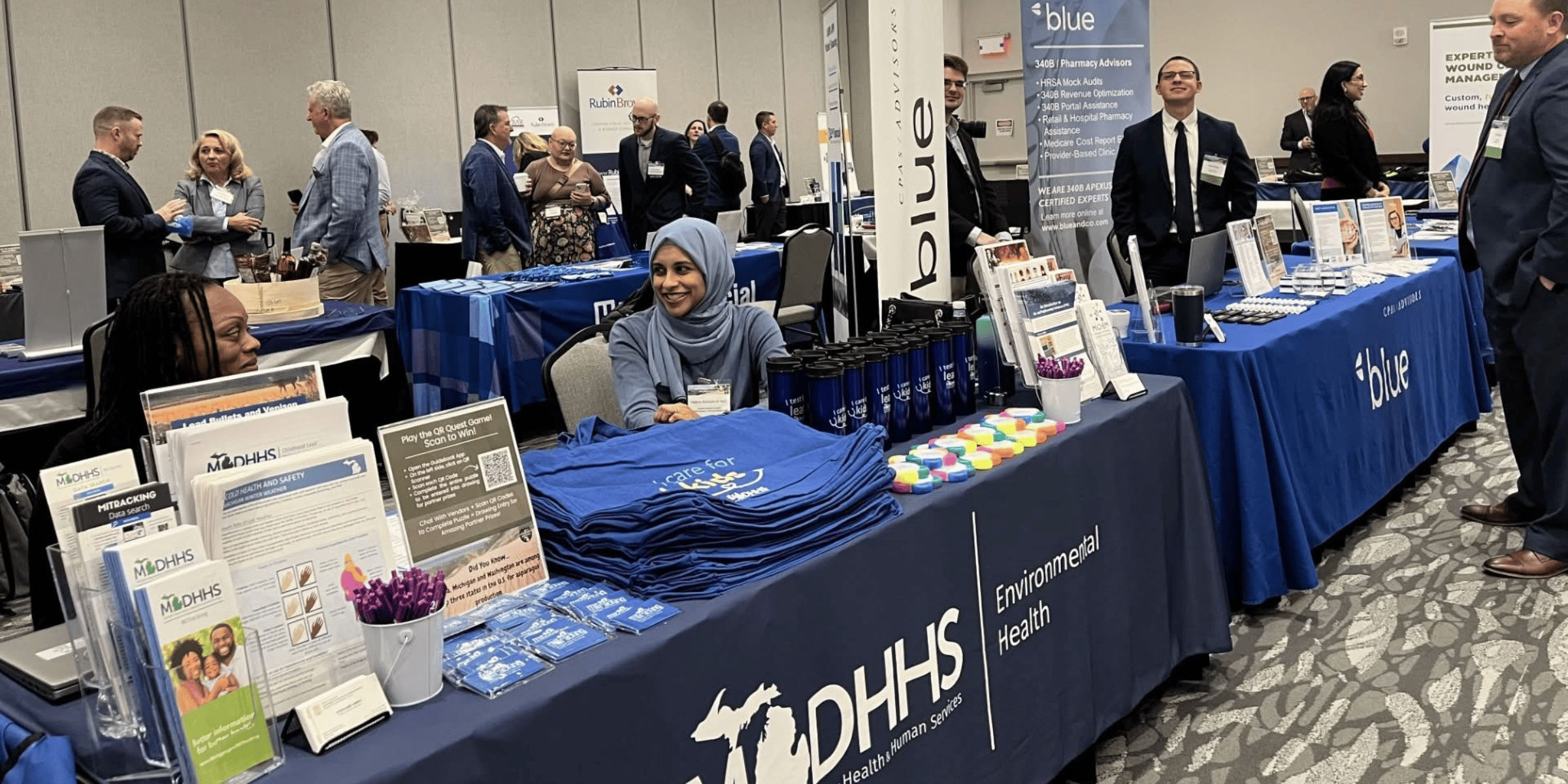 People interacting at exhibition booths with MDHHS and blue promotional materials.
