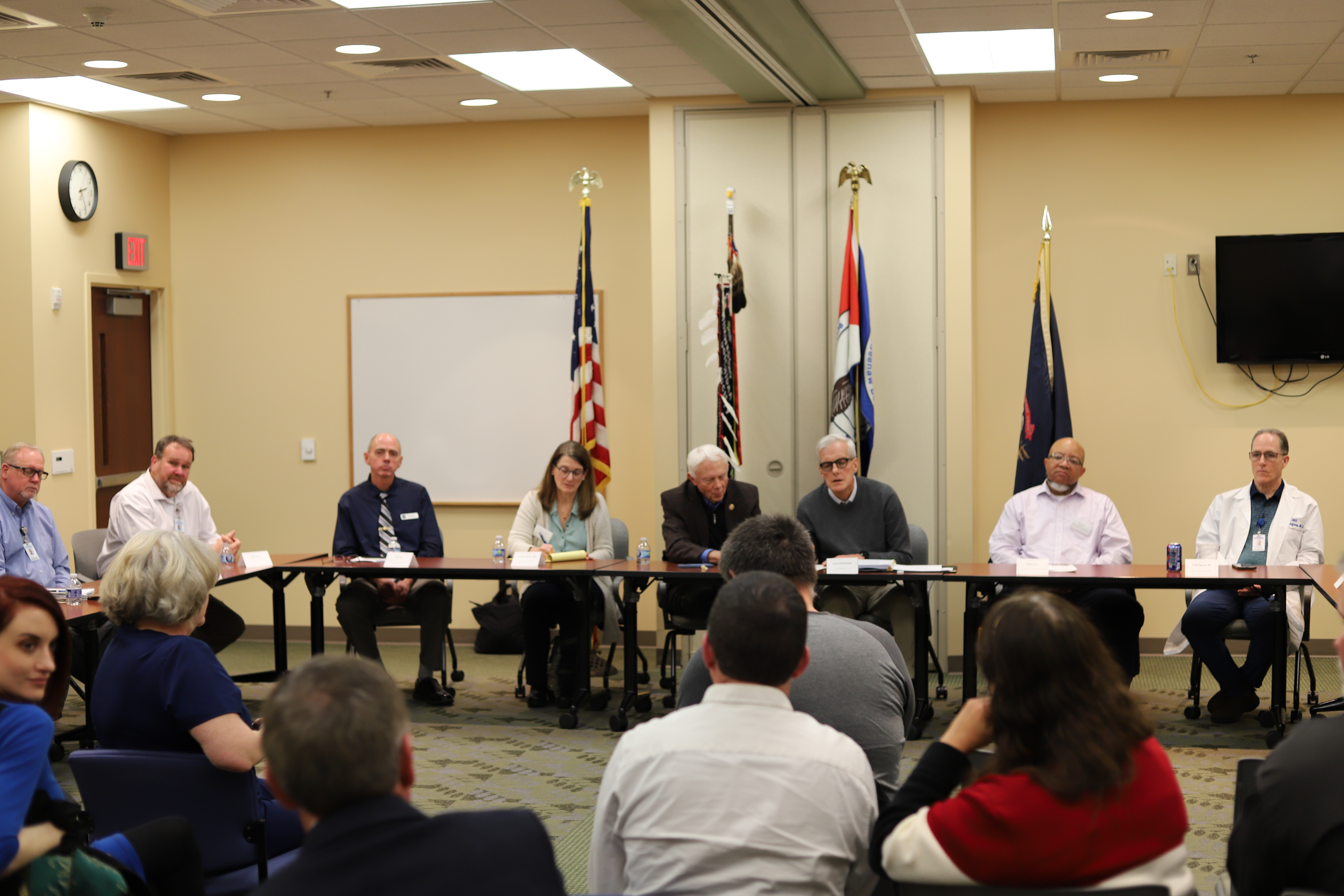 A group of people seated at a conference table in a meeting room with flags and a whiteboard in the background.