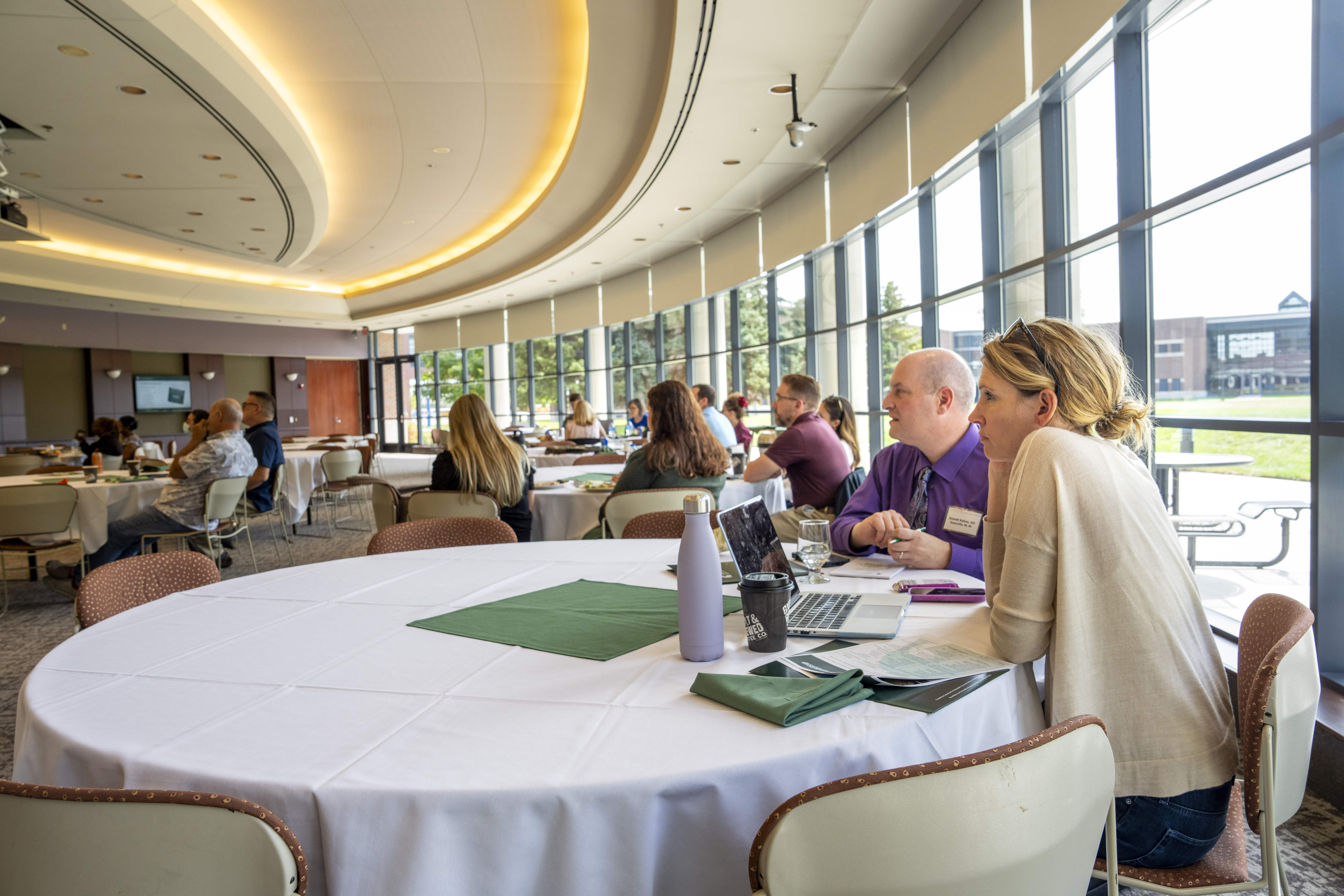 Attendees seated at round tables in a bright, modern conference room with large windows during a daytime event.