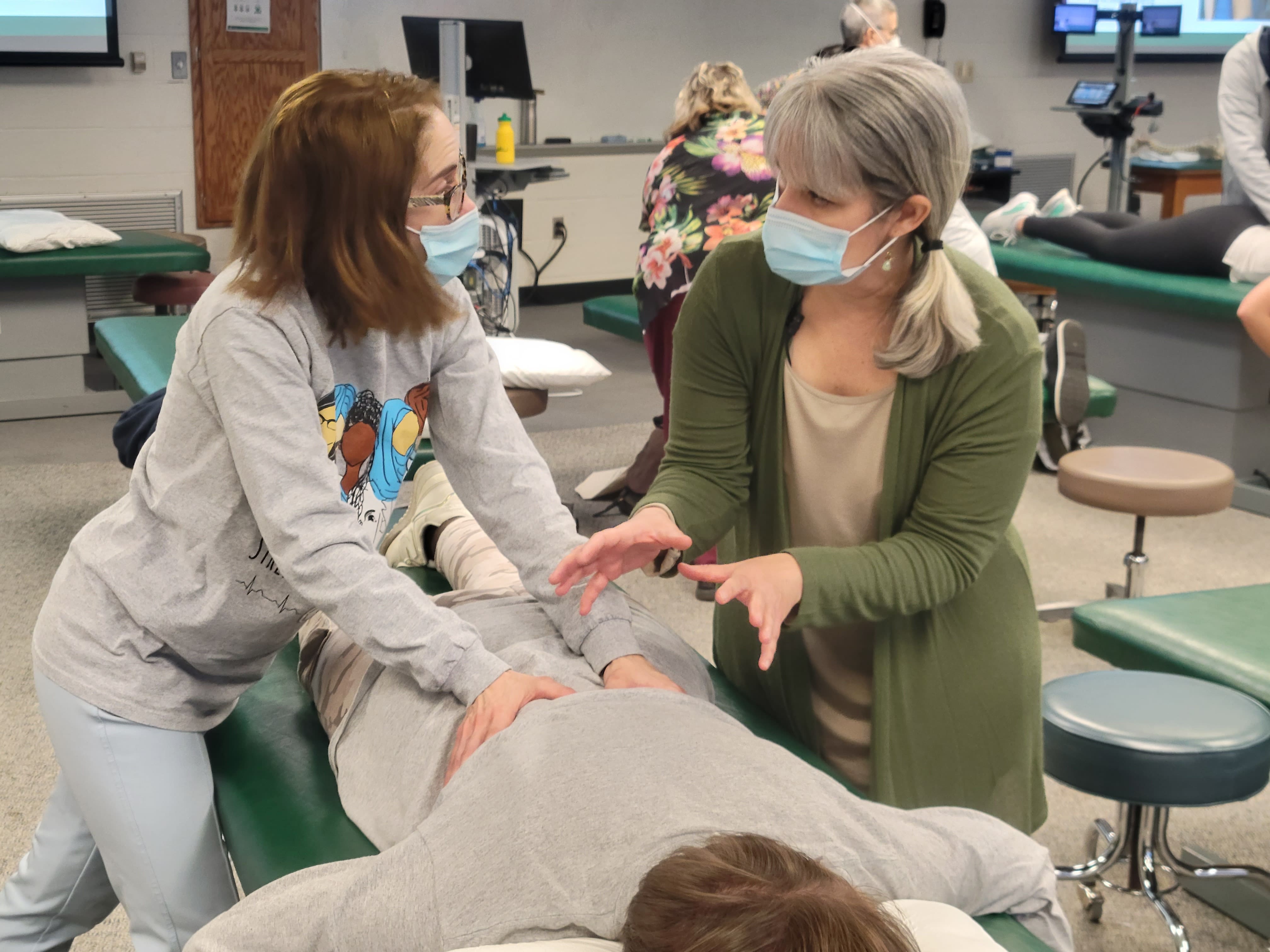 Two people practicing a technique on a person lying on a table in a classroom setting.