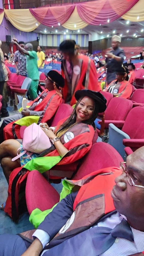 A photo of Dr. Nwando Achebe sitting in an auditorium with other people. She is dressed in a red robe, sitting and has a black hat on her head.