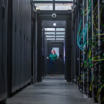 A photo of a person in a green shirt walking in data center, a dark hallway with wires