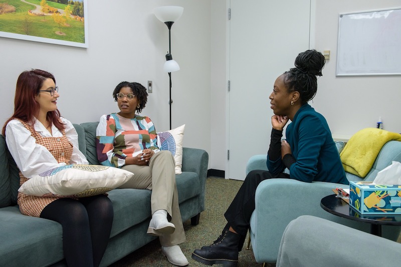Lekie Dwanyen (right) hosts a supervision meeting with CFT student therapists. All three are seated on blue furniture and are talking.