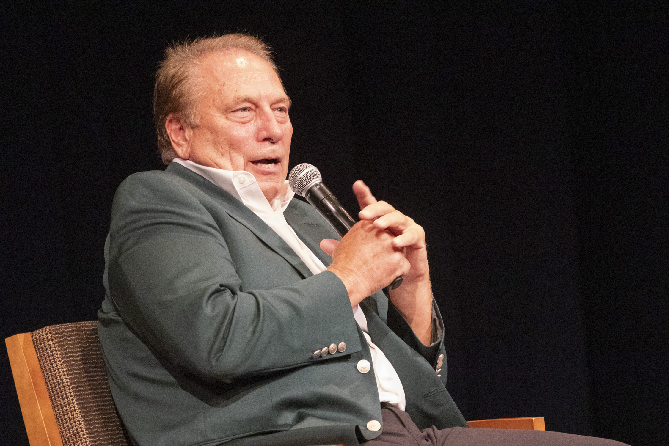 Tom Izzo siting and holding a microphone as he speaks to an audience