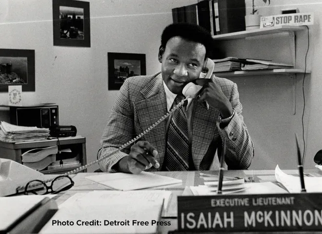 A black and white photo of a man on a phone with a cord sitting at a desk with a sign that says executive lieutenant Isaiah McKinnon with text on the photo that says Photo Credit: Detroit Free press