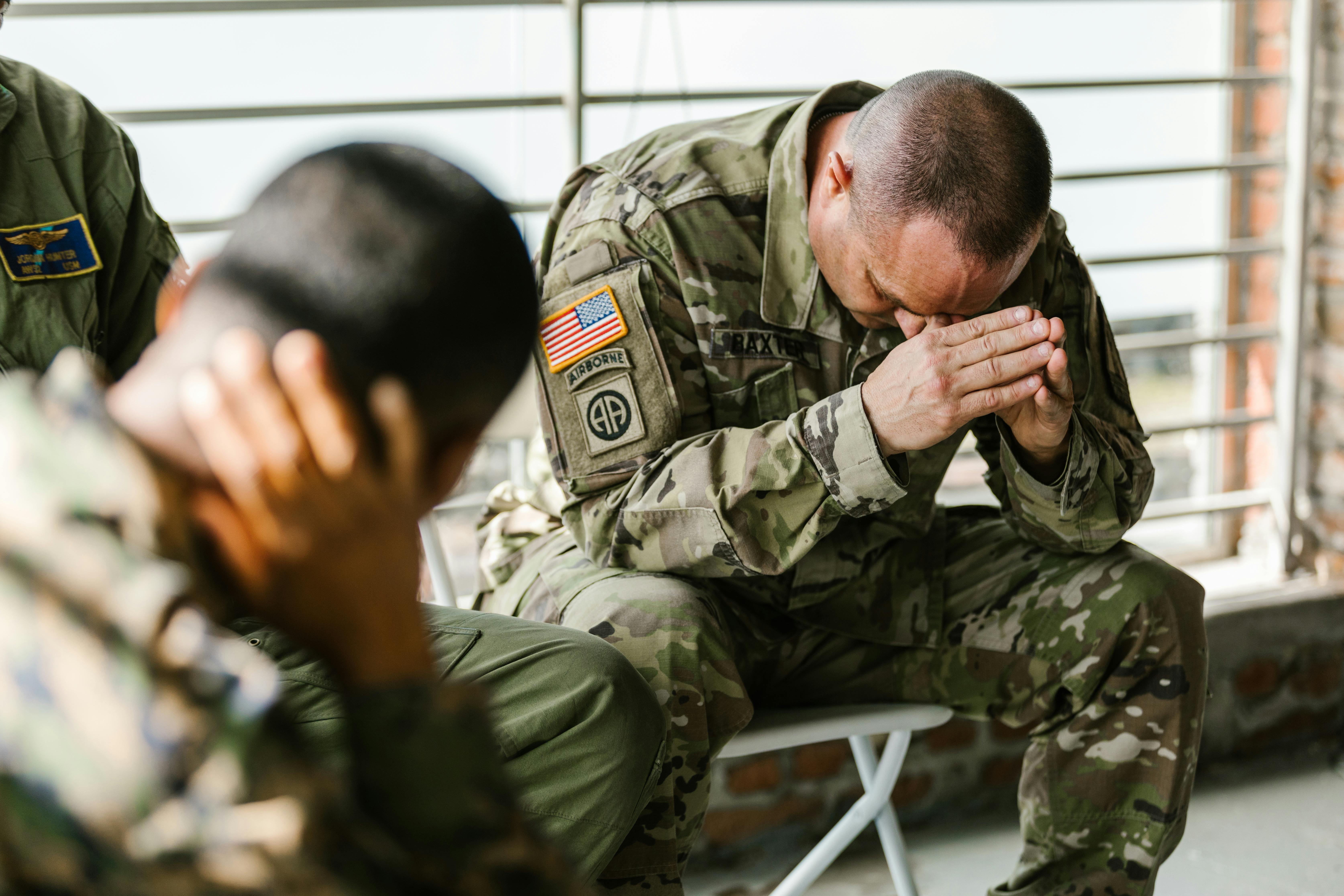 A man in Army fatigues bows over his knees while sitting in a prayer like position