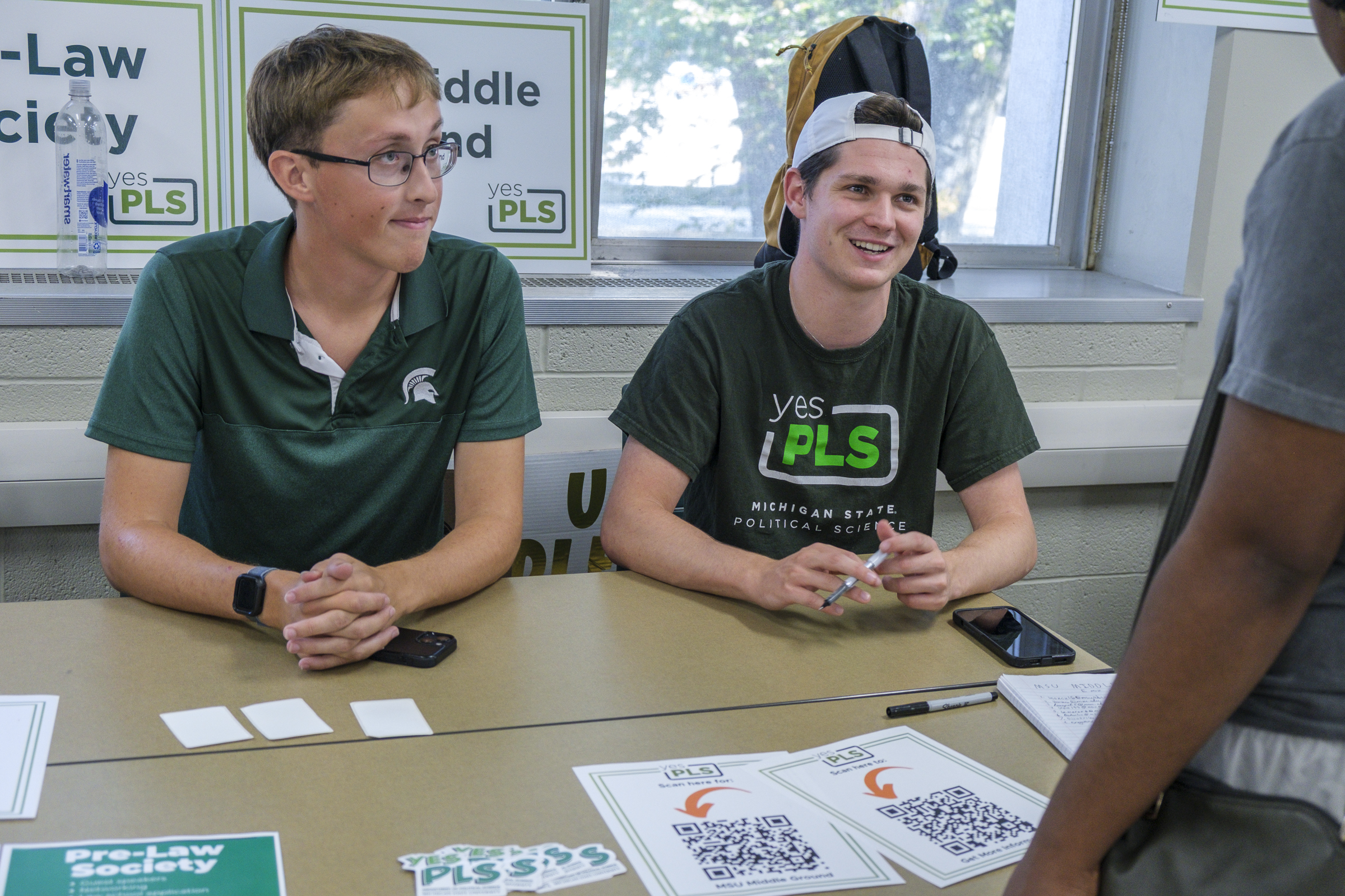 A photo of a student in a white hat on backwards in a green shirt sitting at a table with a sign behind that reads MSU Middle Ground as he sits with another student at the table who has glasses on