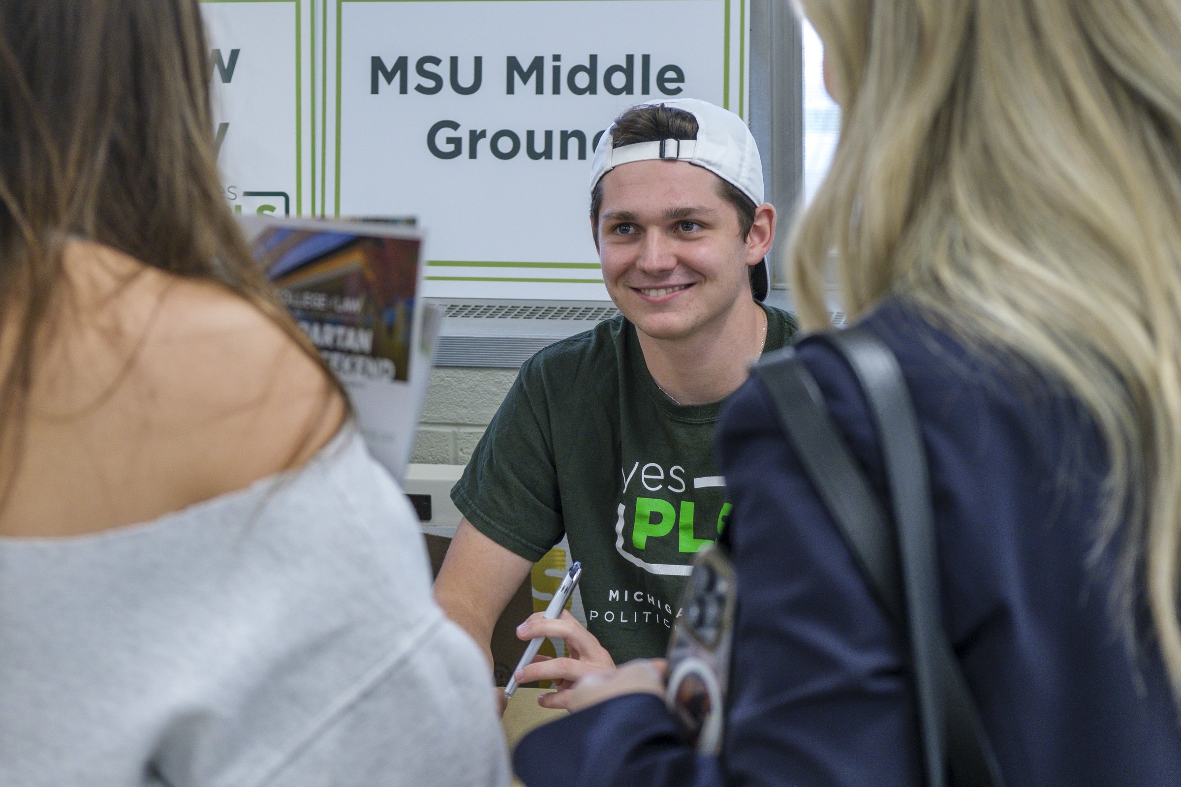 A photo of a student in a white hat on backwards in a green shirt sitting at a table with a sign behind that reads MSU Middle Ground as he talks to people at the table