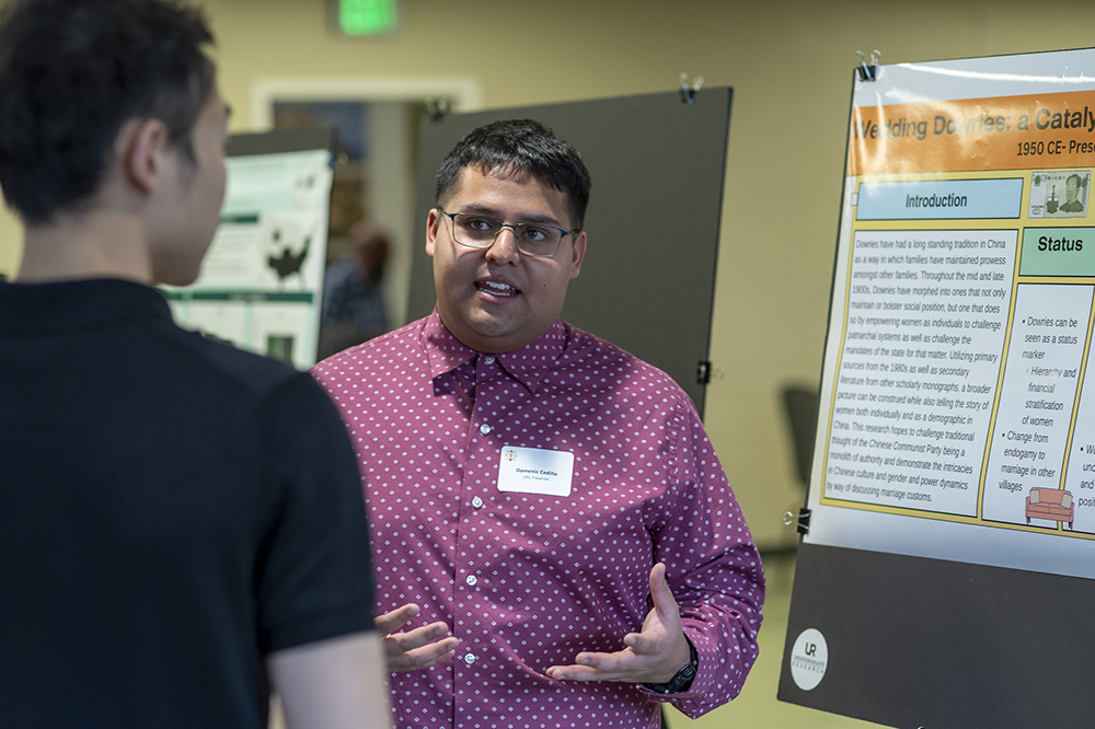 A student stands in front of a poster and talks to another person about his research on the poster