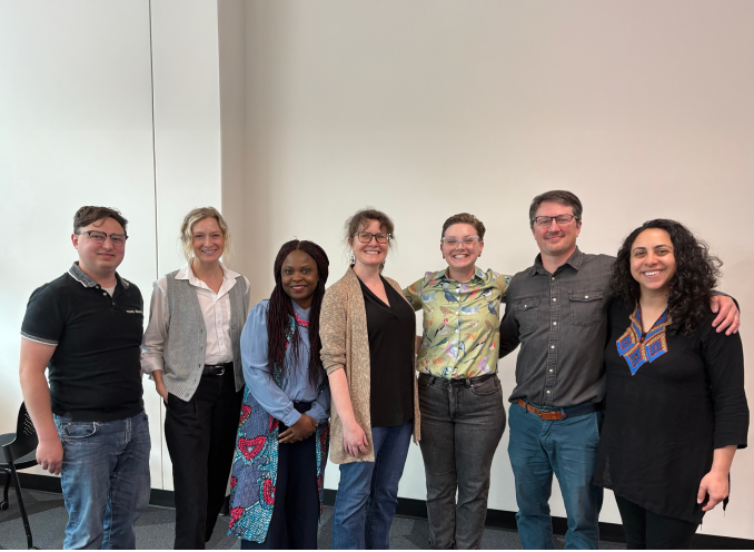 Seven individuals standing in a row indoors, smiling at the camera against a plain light wall.