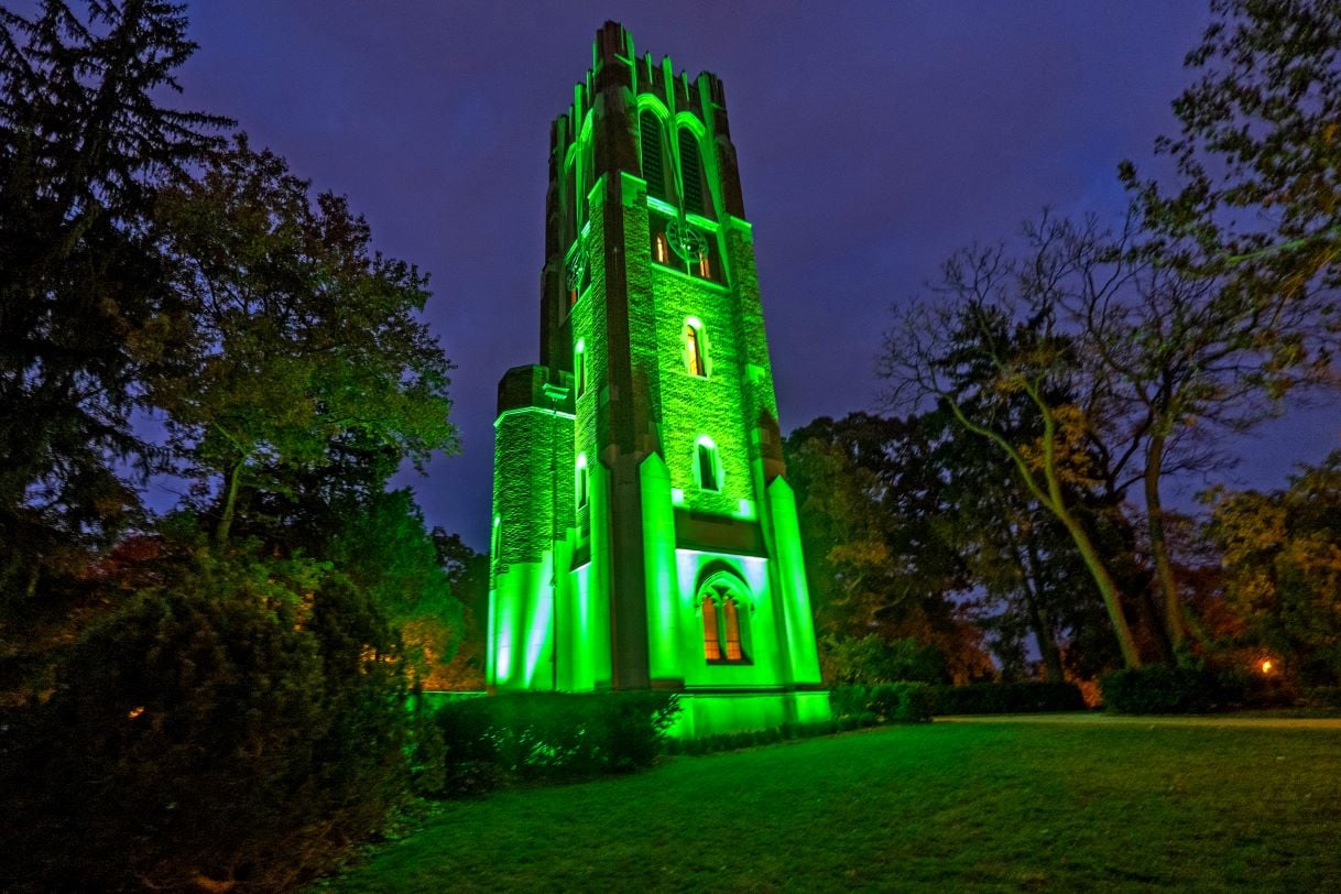 Beaumont Tower at Michigan State University glows green against the night sky, surrounded by trees and illuminated pathways, symbolizing Spartan pride.