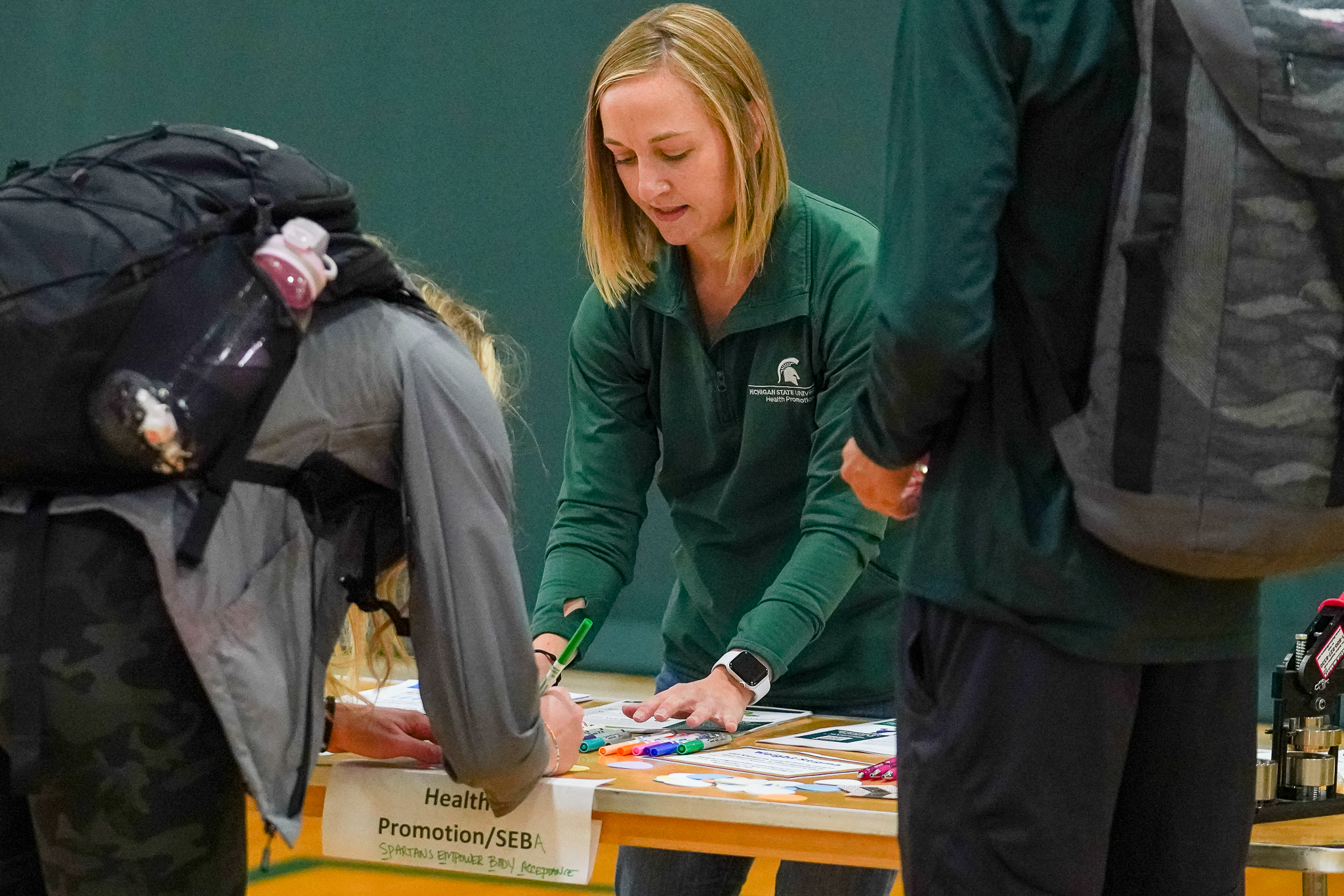 A woman wearing Michigan State University attire helps a man complete an activity at a table.