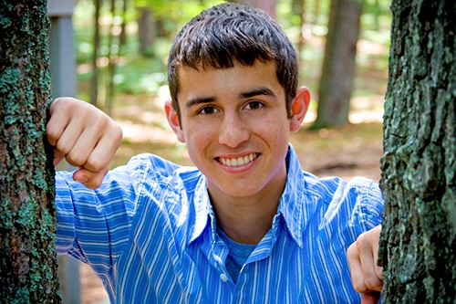Alex Powell standing between two trees, smiling as he leans both arms on the trunks, wearing a striped blue shirt.
