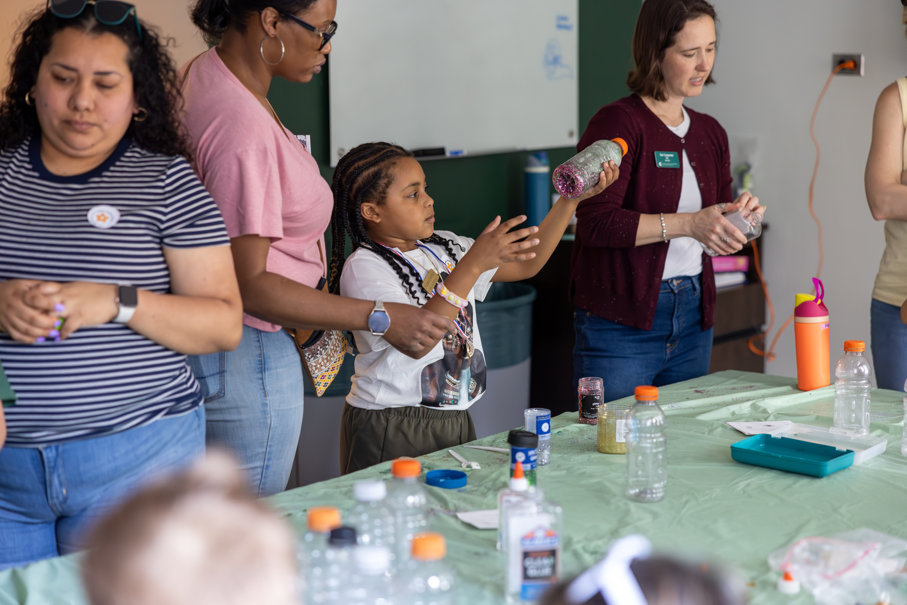 Children and adults stand around a table covered with craft supplies, ready to make calming glitter bottles, which can be shaken to be filled with glitter, and illustrate mindfulness as the glitter settles down and separates with glue and water.