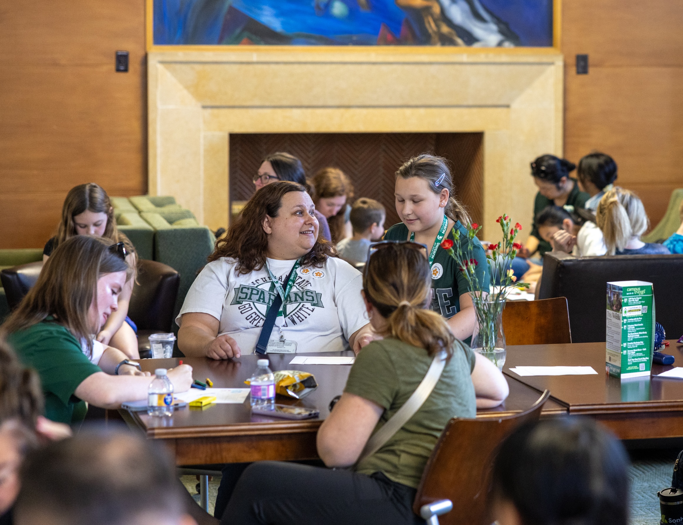 Several children and adults sit around a large table indoors, writing or drawing on paper.