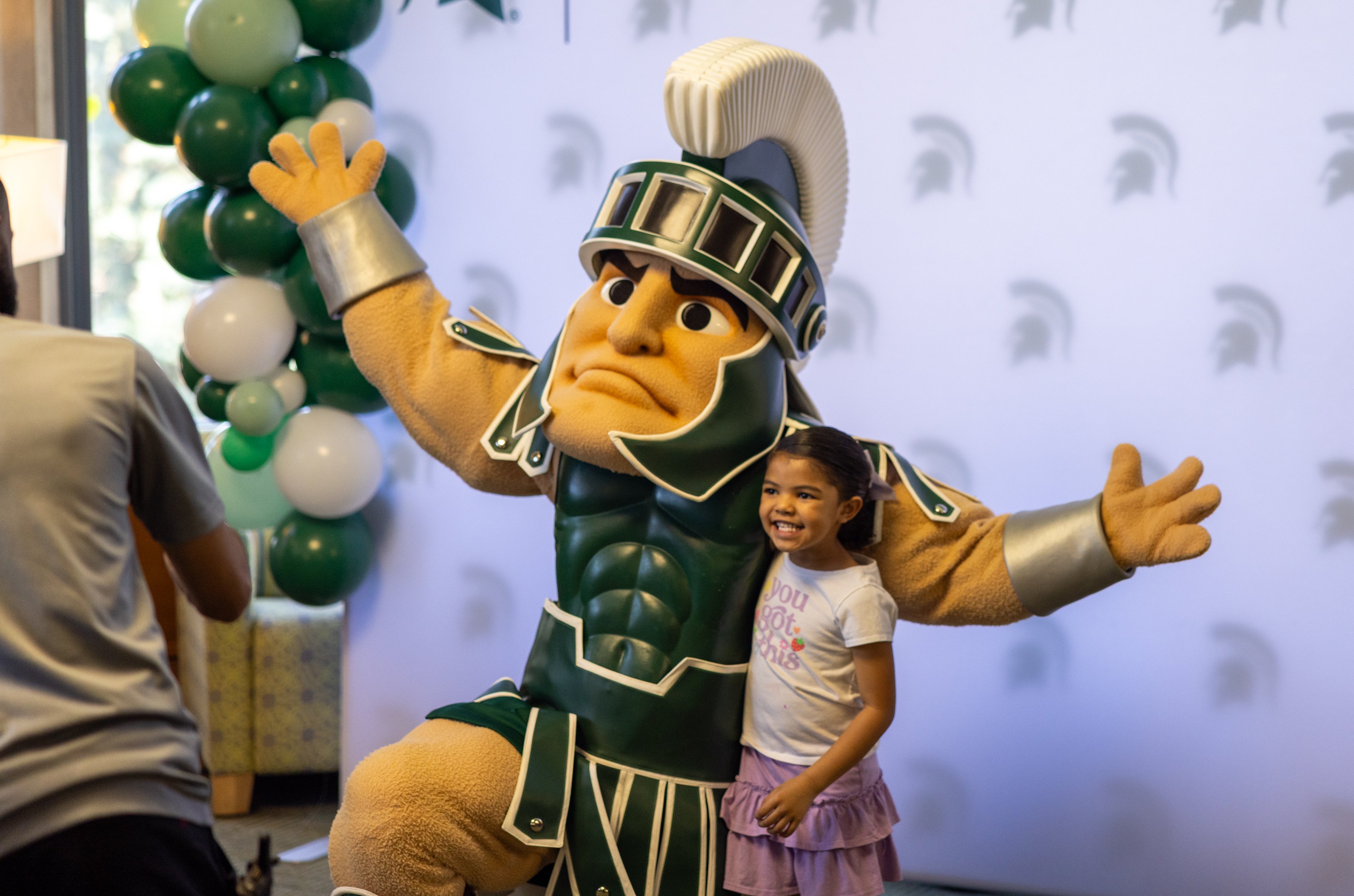 Michigan State University’s Sparty mascot poses with a child in front of a backdrop featuring Spartan helmet logos and green and white balloons.