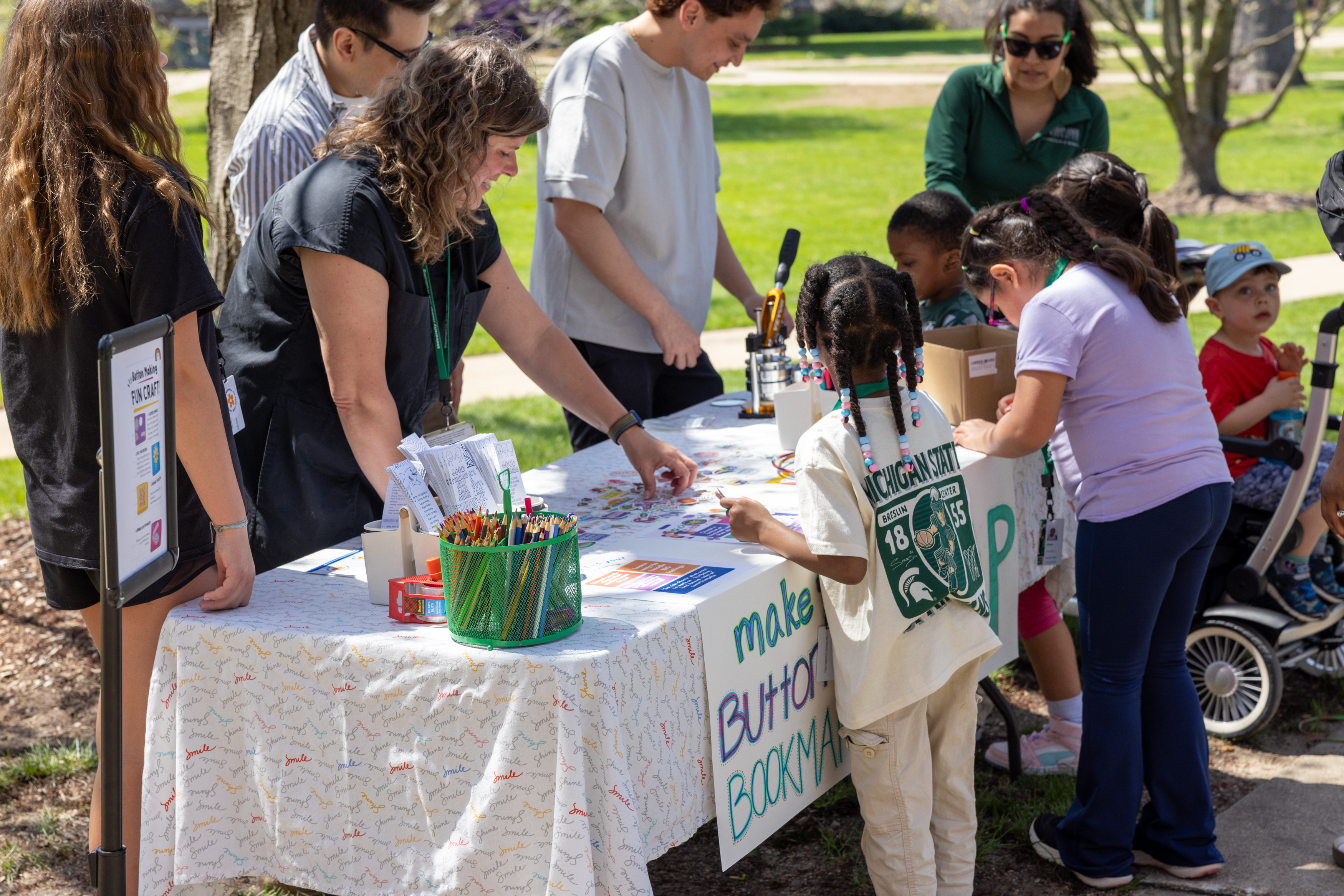 Children and adults gather around an outdoor table labeled “Make a Button Bookmark,” selecting materials from baskets of colored pencils and paper. Staff assist at the table.