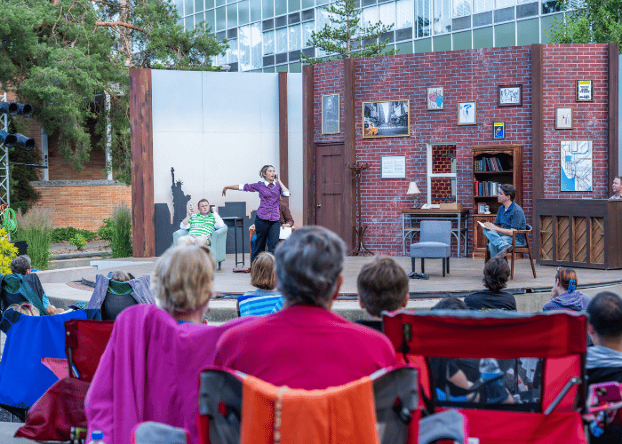 View of a stage from behind a crowd of onlookers. Actor acts out a phone call on the stage.